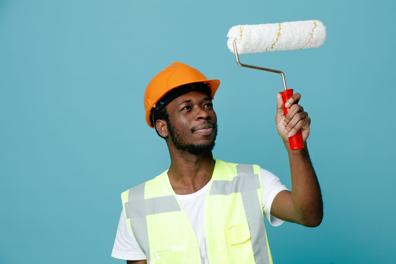 impressed young african american builder in uniform holding roller brush isolated on blue background